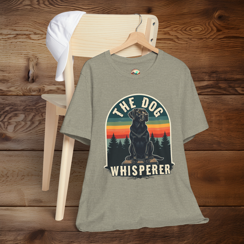 Heather stone (beige-grey) T-shirt featuring a vintage design of a black Labrador retriever on a platform, framed by evergreen trees and a multicolored retro sunset. Text says “The Dog Whisperer.” Shirt is hung on a wooden hanger draped over a chair with a white cap on the backrest.
