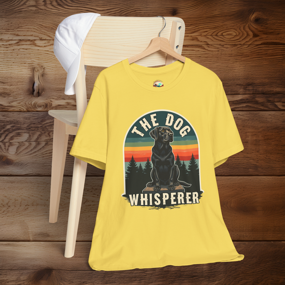 Bright maize yellow T-shirt with a bold retro graphic of a black Labrador against a striped orange, red, and teal sunset background with pine trees. Text reads “The Dog Whisperer.” Displayed on a hanger over a light wooden chair with a white cap on top.