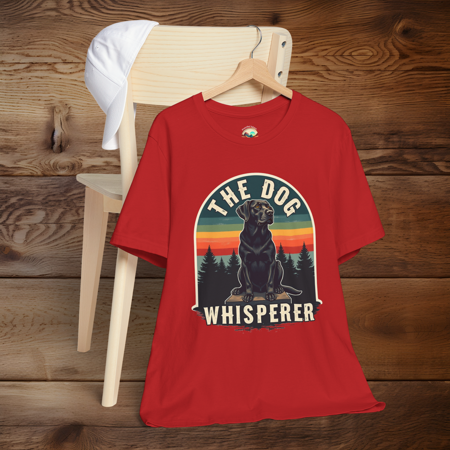 Vibrant red T-shirt featuring a retro design of a black Labrador sitting on a wooden platform, with silhouetted trees and sunset stripes in the background. Text above and below reads “The Dog Whisperer.” Displayed hanging on a wooden chair with a white cap draped on the back.
