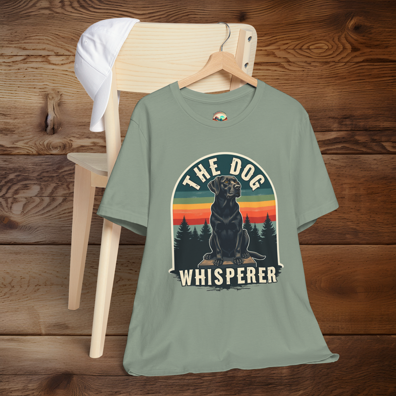 Sage green T-shirt with a rustic retro illustration of a black Labrador in front of pine trees and a multicolored sunset. The arched design includes the words “The Dog Whisperer.” Shirt is shown on a wooden hanger over a chair with a white cap placed on the backrest.