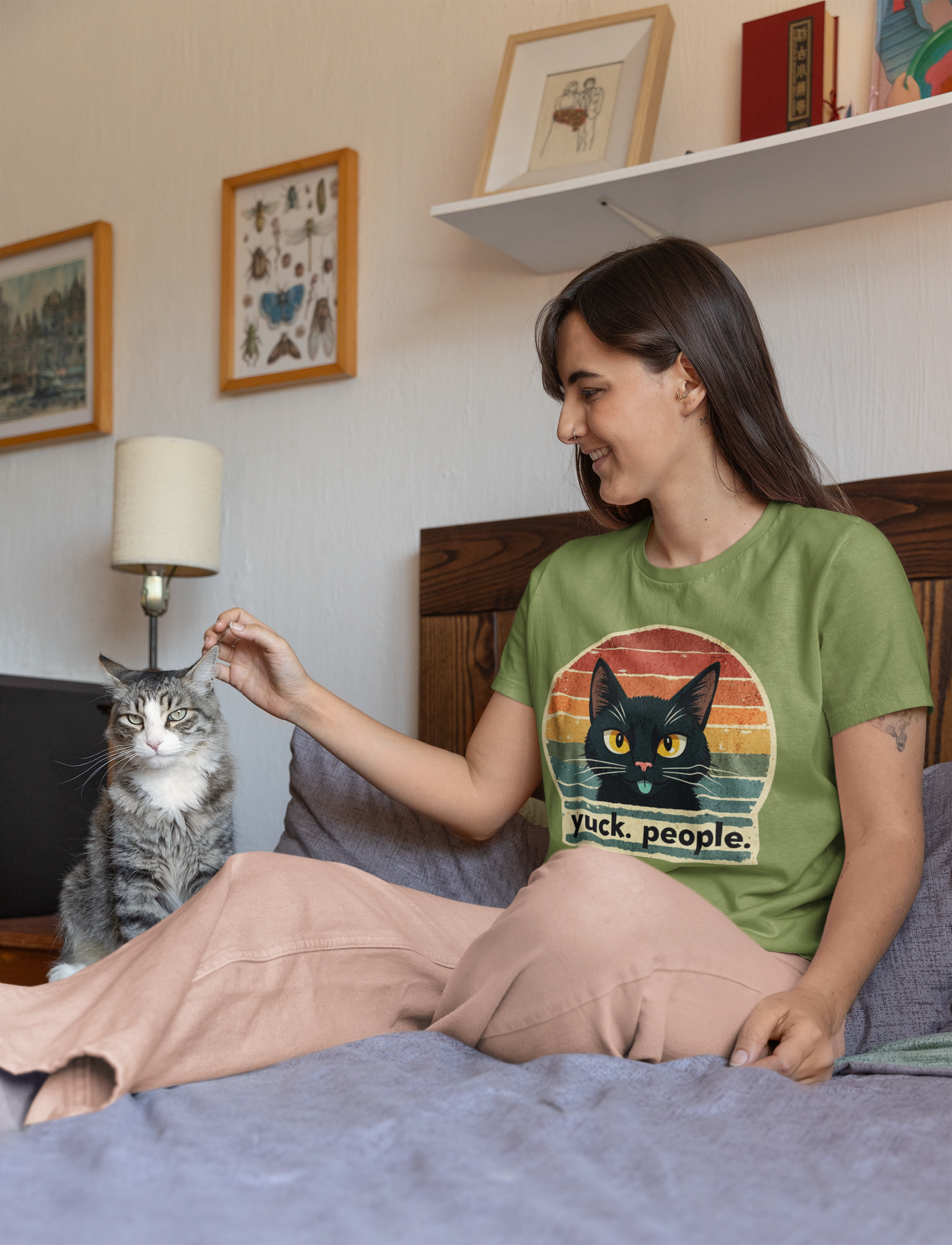 Smiling woman wearing a green “Yuck. People.” retro cat t-shirt while sitting on a bed and petting her tabby cat in a cozy bedroom setting with framed art and books in the background.